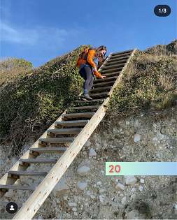 A white female with long brown hair smiles walks up a wooden ladder from the beach